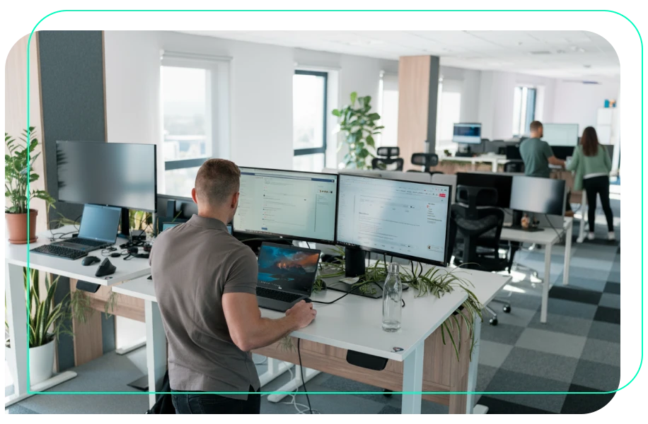 Modern open office space with large windows and natural light. A man in the foreground stands at a white desk with three monitors and a laptop, working on email and web content. The desk is decorated with green plants and a water bottle. In the background, other employees are seen collaborating and working at similar desks. The office has a clean, minimal design with checkered carpet, ergonomic chairs, and tech setups, creating a focused and professional environment.